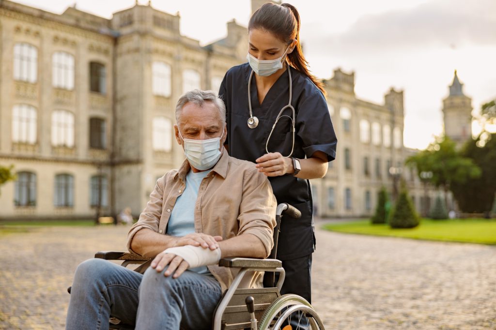 Young nurse in protective mask taking care of senior recovering patient in wheelchair with bandaged