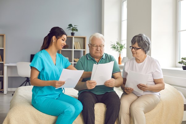 A caregiver discusses documents with two older adults seated on a bed, representing informed decision-making and growing interest in senior care industry investment opportunities.