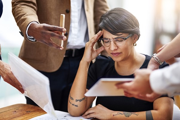 A stressed professional surrounded by documents and colleagues holds her head in frustration, symbolizing corporate burnout and the appeal of transitioning to a fulfilling home care franchise career.