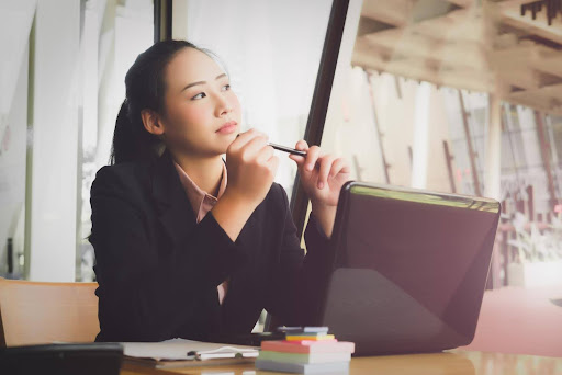 A thoughtful woman in business attire sits at her laptop, holding a pen and looking contemplative, representing the decision-making process between launching a franchise or an independent home care business.