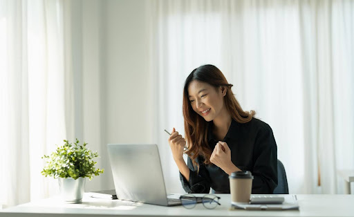 A smiling woman celebrates in front of her laptop at a bright desk, symbolizing early financial wins and promising profit margins for new home care franchise owners in their first year.