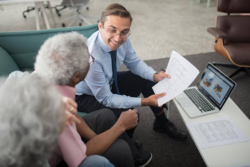 A professional advisor enthusiastically shares documents with an older couple while pointing to a business plan on a laptop, symbolizing the supportive framework available for launching a smart, successful home care business.