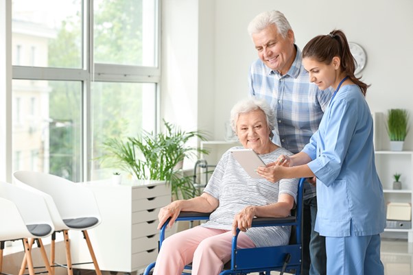 A caregiver shows a tablet to a smiling elderly woman in a wheelchair and her companion, symbolizing the importance of informed decision-making before investing in a home care franchise.