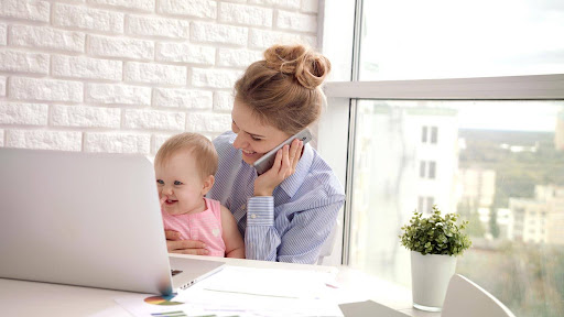 A parent works from home while holding their baby, multitasking between a phone call and a laptop at a bright desk by the window.