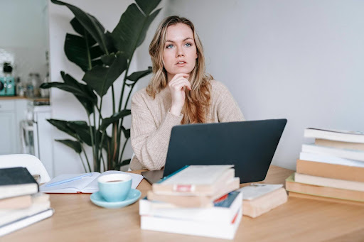 A thoughtful woman sits at a desk with a laptop and stacks of books, representing someone considering if they need to be a business person to succeed in a home care franchise.