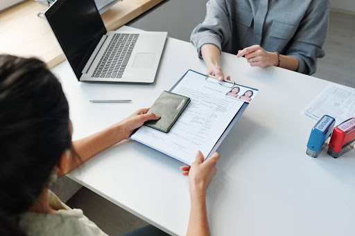 Two women at a desk exchanging a completed immigration form and a passport—illustrating the E-2 visa application process tied to starting a home care business.