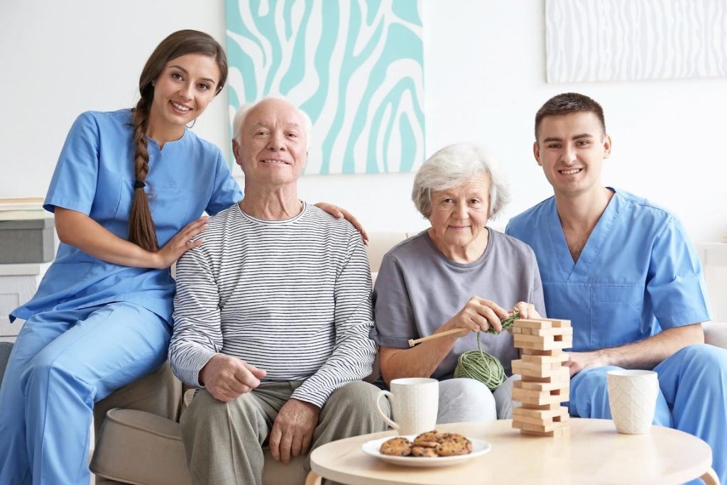 Two cheerful caregivers in scrubs smiling alongside an elderly couple enjoying leisure time together at home, symbolizing the supportive and friendly environment of a successful home care agency.