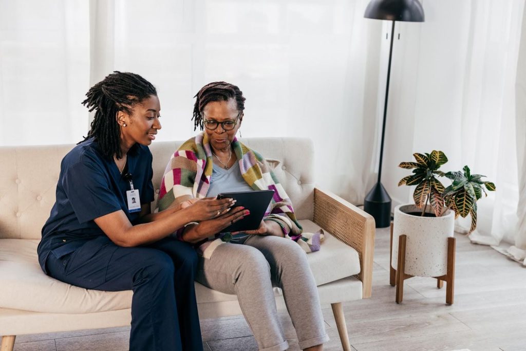 A professional caregiver sits beside an elderly woman wrapped in a colorful blanket, showing her something on a digital tablet. They are seated on a cozy cream-colored couch in a bright, warmly lit living room with potted plants and modern decor.