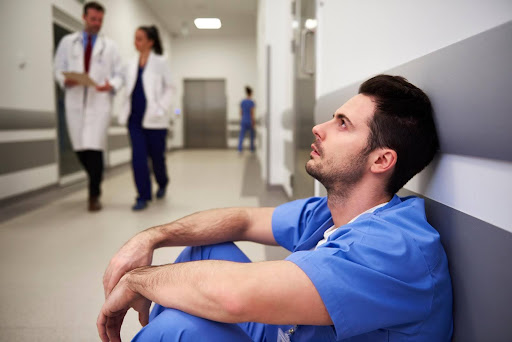 A tired male nurse in blue scrubs sits on the floor in a hospital hallway, looking up with exhaustion, while two medical professionals walk by in the background—representing burnout and underpayment in healthcare.
