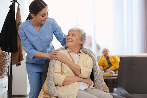 A young female caregiver in blue scrubs gently places a warm blanket over the shoulders of an elderly woman seated on a couch, both smiling warmly at each other, with another senior woman sitting in the background.