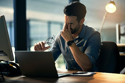 A stressed man sits at his desk with his eyes closed and pinching the bridge of his nose, holding his glasses in one hand, surrounded by paperwork and a laptop—showing classic signs of burnout and job dissatisfaction.