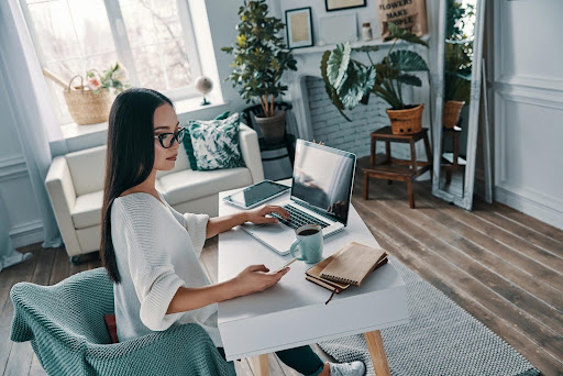 Woman working from home at a stylish desk setup with a laptop, notebook, and coffee—illustrating the possibility of running a home care franchise remotely.