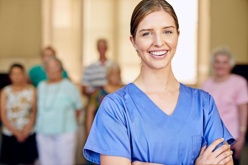 Confident caregiver smiling with arms crossed in front of a group of seniors, representing a franchise opportunity for caregivers aspiring to be CEOs.