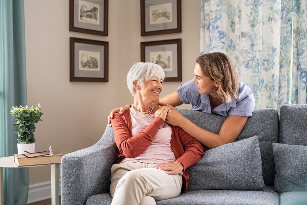 Smiling adult daughter sitting beside her elderly mother on a couch, sharing a warm moment—symbolizing a proud parent inspired by her child's meaningful business.
