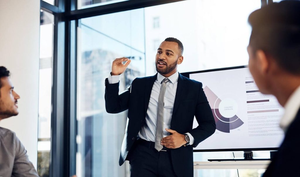 Confident businessman in a suit presenting profit margin data on a screen to colleagues in a modern office setting, comparing franchise and startup home care models.