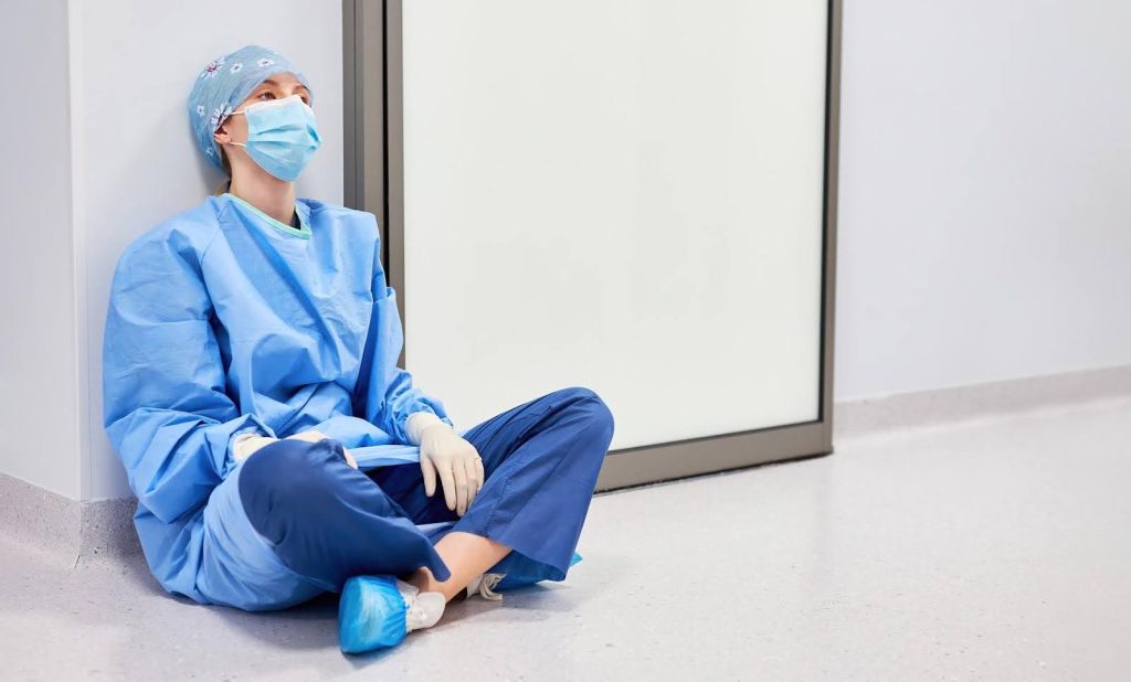 Burn out healthcare worker in scrubs sitting on the floor of a hospital hallway, leaning against the wall with a mask on and eyes looking up in fatigue.