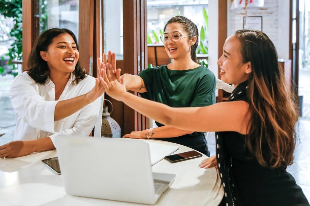 Three smiling women sitting around a table with a laptop, giving each other high-fives, symbolizing teamwork, encouragement, and ongoing support for franchisees after launch.