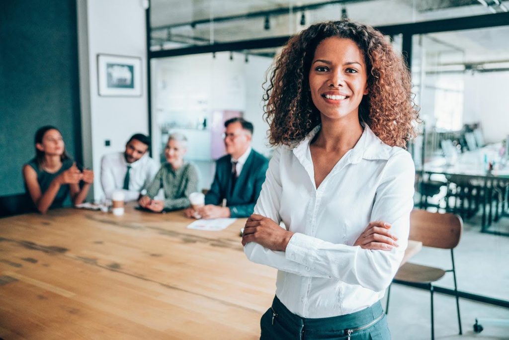 Confident businesswoman standing at the forefront of a meeting room with a team seated behind her, symbolizing leadership traits that contribute to success and growth as a senior care franchisee.