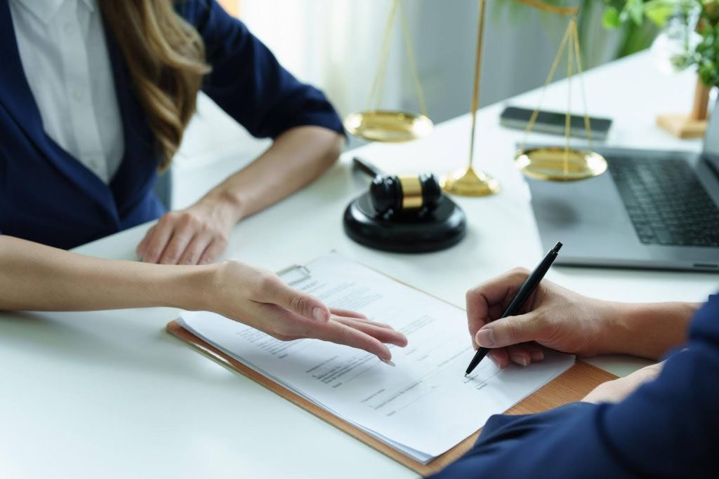 Close-up of a person signing legal documents with a lawyer, with a gavel and scales of justice on the desk, symbolizing the legal requirements for opening a home care franchise in the U.S.