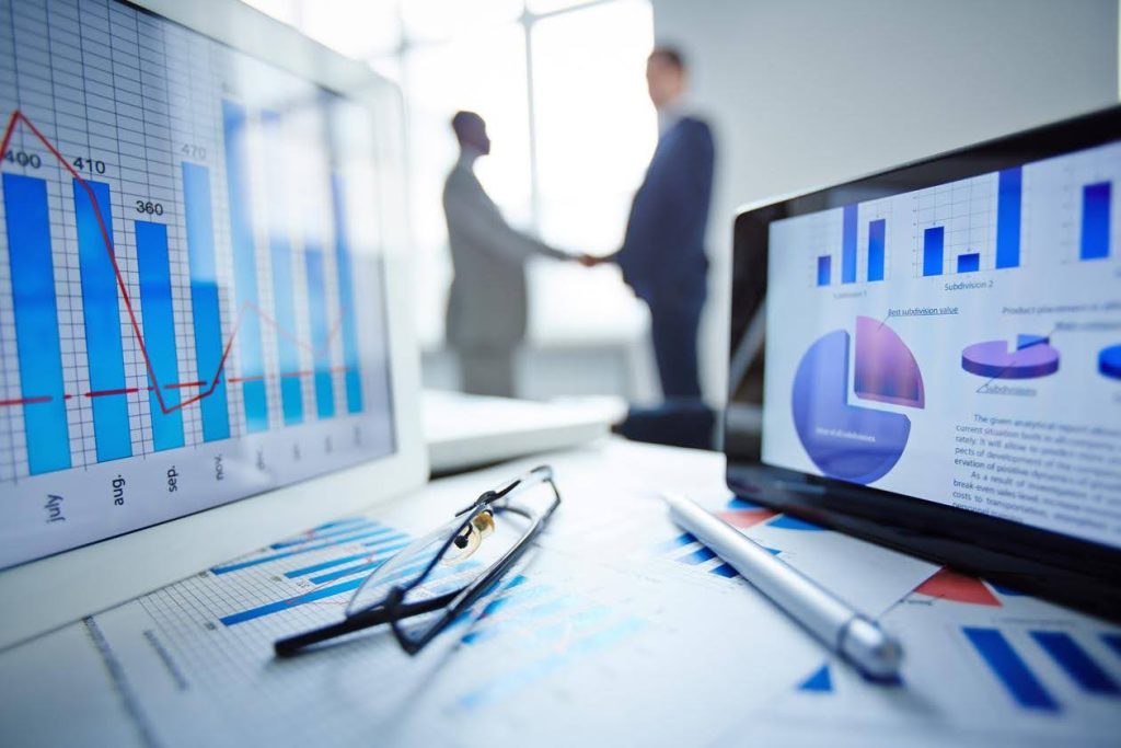 Close-up of financial charts on laptop screens with two business professionals shaking hands in the background, symbolizing a strategic approach to market a home care franchise.
