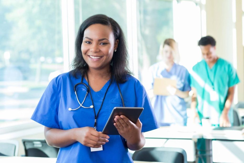 Smiling healthcare professional in blue scrubs holding a tablet with a stethoscope around her neck, symbolizing questions about whether a medical background is required to own a home care franchise.