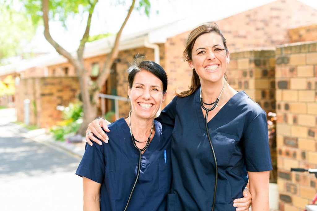 Two smiling female caregivers in navy scrubs standing outdoors with stethoscopes around their necks, representing efforts to recruit and train quality caregiving staff as a franchise owner.