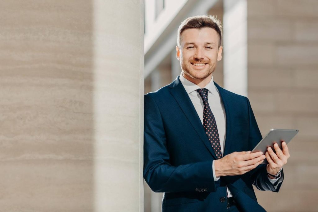 Confident businessman in a suit holding a tablet and smiling, representing the traits of a successful franchisee who stands out from others.