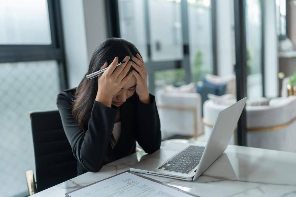 Tired employee sitting at a desk with a laptop, holding her head in frustration, representing burnout and the desire for a new career path.