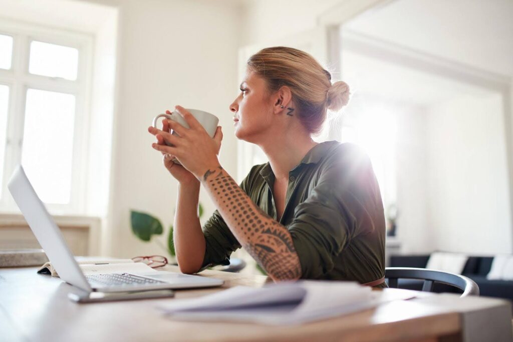 Thoughtful woman sitting at a desk with a laptop and coffee cup, symbolizing someone considering whether to start a home care franchise or run an independent business.