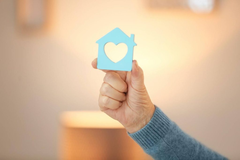 Close-up of a senior hand holding a small blue house cutout with a heart shape, symbolizing the stability and compassion behind home care businesses that remain strong even during a recession.