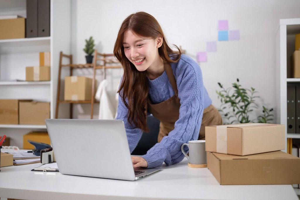 Smiling woman working on a laptop surrounded by packages, symbolizing running a home care franchise remotely.